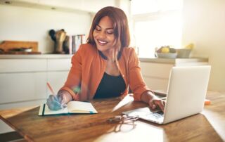 A woman smiles while taking notes from a laptop screen in a notepad.